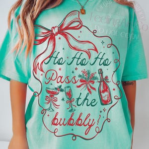 May include: A turquoise t-shirt with a festive design. The graphic features a red bow, the words "Ho Ho Ho Pass the bubbly!" in red and green, and illustrations of champagne glasses and a bottle.