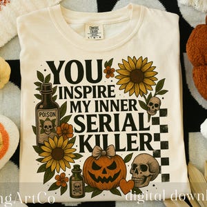 May include: Cream-colored t-shirt with the text "YOU INSPIRE MY INNER SERIAL KILLER" in black, with floral and skull graphics. The shirt is surrounded by Halloween-themed decorations, including pumpkins and ghost-shaped ornaments.