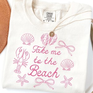 May include: Cream-colored t-shirt with pink illustrations of seashells, a lobster, starfish, and bows. The text "Take me to the Beach" is written in a cursive font. A gold necklace and a brown hat are also visible.