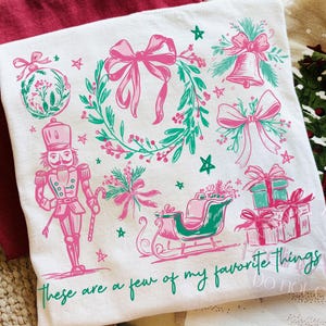 May include: White t-shirt with a pink and green Christmas design. The design includes a wreath, bows, a bell, a nutcracker, a sleigh, and gifts. The text "These are a few of my favorite things" is printed at the bottom.