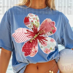 May include: A light blue t-shirt featuring a large, appliqued flower design with red, pink, and white fabric patches. The flower has a detailed, stitched center. The shirt is worn near the ocean.