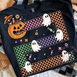 May include: Black t-shirt featuring a Halloween design. The graphic includes a jack-o'-lantern, ghosts, and candy. The design incorporates checkered patterns in orange, pink, green, and purple. The shirt is displayed on a neutral background.