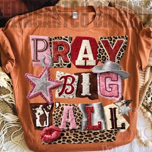 Può includere: T-shirt arancione con la scritta "PRAY BIG Y'ALL" in stile patchwork. Il design include un ferro di cavallo, una stella, un cappello da cowboy e labbra. Le lettere hanno varie trame, tra cui stampa leopardata e glitter.