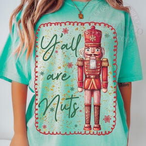 May include: A turquoise t-shirt featuring a festive design. The design includes a red-coated nutcracker soldier with the words "Y'all are Nuts" in green script. The design is framed with a red border and snowflake accents.