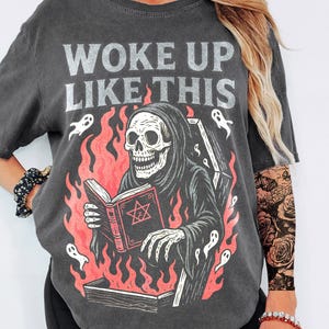 May include: Dark grey t-shirt with the text "WOKE UP LIKE THIS". The graphic features a skeleton in a hooded robe reading a book with a pentagram, surrounded by flames and ghosts. A gothic-themed tee.