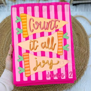 May include: A rectangular fabric item with a bright pink and light pink vertical striped background. It features three horizontal carrot-shaped graphics with orange striped tops and light orange bodies. The text reads Count it all joy James 1:2.