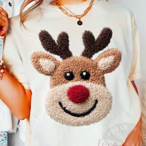 以下が含まれることがあります： 赤い鼻、茶色の角、笑顔の口を持つトナカイの顔が特徴のクリーム色のTシャツ。トナカイのデザインは、質感のある豪華な外観をしています。「Original Designer Feeling Do not copy」というテキストがシャツにプリントされています。