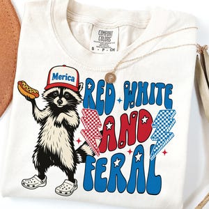 May include: A white t-shirt with a cartoon raccoon wearing a red, white, and blue "Merica" hat and holding a hot dog. The shirt has the text "Red White and Feral" in a retro font.