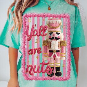 May include: A turquoise t-shirt featuring a pink and white striped rectangle with the words "Y'all are nuts" and a pink nutcracker design. The nutcracker has a black mustache, a white beard, and a pink hat.