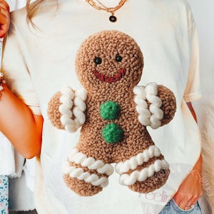 May include: A cream-colored t-shirt featuring a brown gingerbread man design with white and green accents. The gingerbread man has a smiling face and is made of a textured material. The shirt is worn with blue jeans.