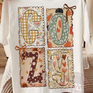 May include: White t-shirt with the word "COZY" in a fall-themed design. The letters are filled with patterns like gingham, floral, and polka dots, with pumpkin and leaf accents. A brown bow is on the top of the letter "O" and "Z".