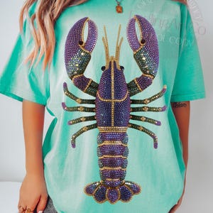 May include: A turquoise t-shirt featuring a large, bejeweled lobster design. The lobster has purple claws, a purple body with gold accents, and green legs. The shirt is a comfortable, oversized fit.