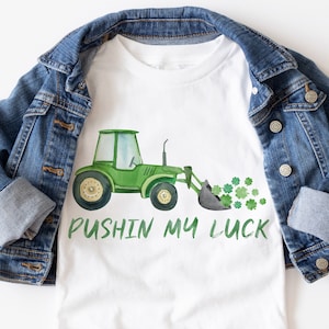 May include: White t-shirt with a green tractor graphic and the text "Pushin My Luck" in green. The tractor is decorated with shamrocks. A denim jacket is draped over the shirt.