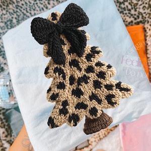May include: A knitted Christmas tree ornament with a leopard print pattern in tan and black. The tree is topped with a large black bow. The ornament is on a light blue surface.