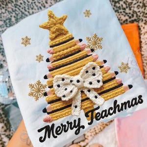 Könnte beinhalten: Hellblaues T-Shirt mit einem Weihnachtsbaum-Design aus gelben Bleistiften, einem goldenen Stern und einer weißen Schleife mit schwarzen Punkten. Der Text "Merry Teachmas" ist schwarz gestickt.
