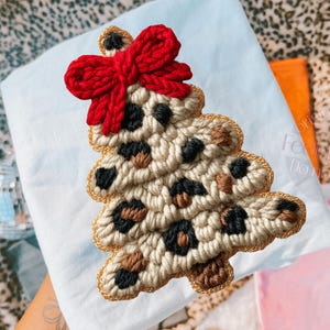 May include: A handmade Christmas tree ornament with a leopard print design. The tree is made of beige yarn with black and brown spots, a gold border, and a red bow at the top. The ornament is on a light blue surface.