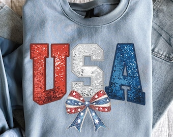 Pode incluir: Uma camisa de frio azul claro com a palavra "USA" em letras de glitter vermelho, branco e azul. Abaixo das letras, há um laço vermelho, branco e azul com estrelas.
