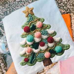 Puede incluir: Diseño bordado de árbol de Navidad con detalles en hilo verde, blanco, rojo, rosa y turquesa. El árbol está coronado con una estrella dorada y se encuentra sobre una tela azul claro. Decoración festiva navideña.