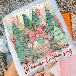 May include: Light blue t-shirt featuring a Christmas-themed illustration. The design includes Christmas trees, a pink bow, birds, and the text "Let heaven & nature sing". The artwork is in shades of green, pink, and gold.