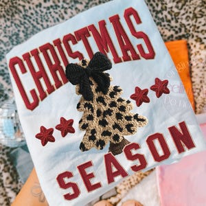 May include: Light blue Christmas-themed t-shirt with the words "CHRISTMAS SEASON" in red block letters. A leopard print Christmas tree with a black bow is embroidered on the shirt, along with three red stars.