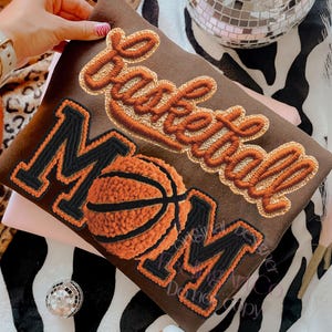 Puede incluir: Una sudadera marrón con las palabras "Basketball Mom" bordadas en naranja y negro. También hay un gráfico de baloncesto bordado en la parte delantera. La sudadera es sostenida por una persona que lleva un reloj blanco.