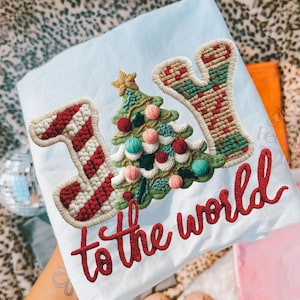May include: Light blue Christmas-themed shirt with the embroidered words "JOY to the world." The design includes a Christmas tree, candy cane, and the letter "Y." The embroidery features red, green, and tan colors.