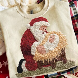 May include: Cream-colored sweatshirt featuring a detailed embroidered design of Santa Claus and baby Jesus in a manger. Santa is in a red suit with a white beard. The baby is in a manger made of straw. A festive holiday-themed garment.