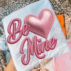 May include: A light blue t-shirt with the embroidered words "Be Mine" in pink script. A pink heart with a scalloped edge is embroidered above the word "Mine". The shirt is folded and resting on a patterned surface.
