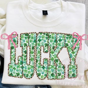 May include: White sweatshirt with the word "LUCKY" in green shamrock and leopard print design. The word "LUCKY" has pink bow accents. The sweatshirt is folded on a white surface with a cup of coffee and a pair of jeans.