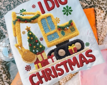 Puede incluir: Camiseta azul claro con temática navideña con el texto "I DIG CHRISTMAS". El diseño presenta una excavadora amarilla decorada con guirnaldas y un árbol de Navidad, junto con cajas de regalo y copos de nieve.