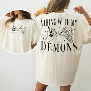 May include: Cream-colored oversized t-shirt featuring a black graphic of two hands making a pinky promise, one human and one skeletal. The back of the shirt has the text "Vibing With My Demons" in a bold, black font, with star accents.