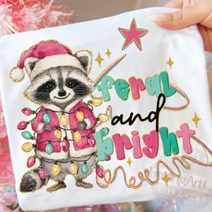 May include: White t-shirt featuring a cartoon raccoon wearing a Santa hat and a pink jacket adorned with Christmas lights. The text "feral and bright" is written in colorful letters with a star and sparkles.