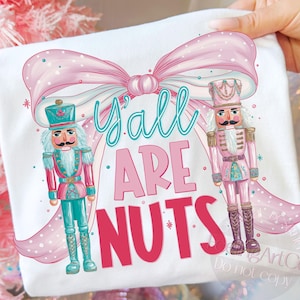 Könnte beinhalten: Weißes T-Shirt mit rosa Schleife und dem Text "Y'all ARE NUTS." Mit zwei Nussknacker-Figuren in Rosa und Türkis. Das Design ist skurril und festlich, perfekt für die Ferienzeit.