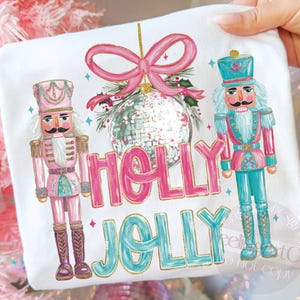 May include: White t-shirt featuring a festive design with two nutcrackers, a disco ball ornament, and a pink bow. The words "HOLLY JOLLY" are written in pink and teal with gold accents. The design has a Christmas theme.