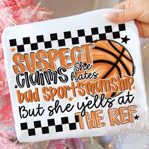 May include: White t-shirt with a basketball graphic and text that reads "SUSPECT Claims she hates bad sportsmanship But she yells at THE REF." The design includes orange and black accents.