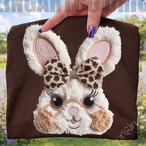 Könnte beinhalten: Ein braunes T-Shirt mit einem Plüsch-Hasengesicht, einer Schleife mit Leopardenmuster, Wimpern und rosa Innenohren. Im Hintergrund ist der Text "FEELINGARTCO ORIGINAL" zu sehen.