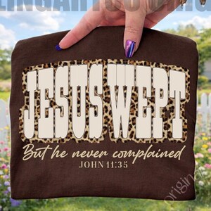 May include: A brown t-shirt with the text "JESUS WEPT" in large, bold letters with a leopard print border. Below, the shirt reads "But he never complained" and "JOHN 11:35" in a smaller, elegant font. The shirt is held up against a background of green grass, a white picket fence, and a blue sky.