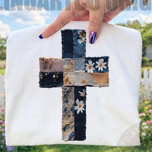 以下が含まれることがあります： パッチワークの十字架のデザインが施された白いスウェットシャツ。十字架は、花柄やデニム柄など様々な生地で作られており、デイジーのアクセントが施されています。背景には白い柵と芝生エリアがあります。