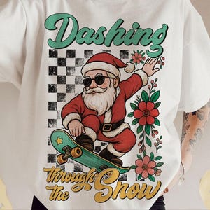May include: White t-shirt with a graphic of Santa Claus skateboarding, wearing sunglasses and a red suit. The text "Dashing Through The Snow" is in a retro font. A black and white checkered pattern is behind Santa.