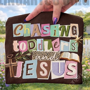 May include: A brown t-shirt with the words "Chasing Toddlers and Jesus" in a collage style. The design includes a cross, an open book, and a star. The background shows a white picket fence and a green lawn.