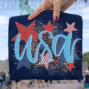 Peut inclure: Un t-shirt bleu marine avec le mot "USA" en écriture bleu clair, entouré d'étoiles rouges et bleues. Les étoiles ont divers motifs, dont des pois et des rayures. L'arrière-plan présente une image floue de la Statue de la Liberté.