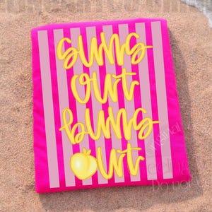 May include: A bright pink beach towel with vertical light pink stripes. The towel features the text "Suns Out Buns Out" in yellow script, along with a yellow peach graphic. The towel is lying on a sandy beach.