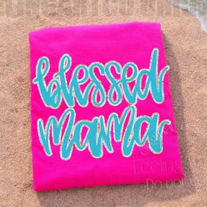 May include: A folded, hot pink t-shirt with the words "Blessed Mama" in a turquoise script font. The shirt is lying on a sandy beach with the ocean in the background. The text has a white outline.