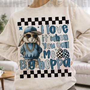 May include: Beige sweatshirt with a graphic of a cool bunny wearing sunglasses, a blue cap, and a gold chain. The text reads "I love it when you call me Big Hoppa" with checkered accents and Easter egg graphics.