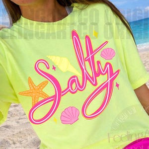 May include: A neon yellow t-shirt with the word "Salty" in pink script, adorned with seashell and starfish graphics. The shirt is paired with bright pink shorts, creating a vibrant beach-themed outfit.