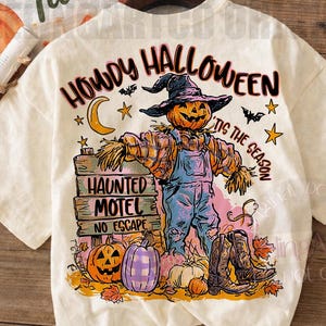 Può includere: Una t-shirt color crema con una stampa grafica di uno spaventapasseri con cappello e salopette, accanto a un cartello in legno con la scritta "HAUNTED MOTEL NO ESCAPE". Il design include zucche, stivali da cowboy e il testo "HOWDY HALLOWEEN 'TIS THE SEASON".