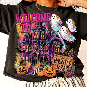 May include: A charcoal gray short-sleeve t-shirt featuring a vibrant graphic print of a haunted mansion, three ghosts, pumpkins, and a sign that reads "WELCOME TO the HAUNTED LIBRARY". The shirt is paired with white and black vertical striped shorts.