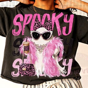May include: A charcoal gray t-shirt featuring a graphic of a ghost wearing a pink leopard print bow, cat-eye sunglasses, and a pink feather boa. The ghost holds a coffee cup and a matching leopard print purse. The word SPOOKY is printed in pink.
