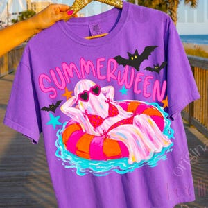 K&ouml;nnte beinhalten: Ein lila T-Shirt mit dem Schriftzug "SUMMERWEEN" in Pink und Gelb. Die Grafik zeigt ein Gespenst mit Sonnenbrille und Bikini, das auf einem bunten Schwimmring treibt. Schwarze Flederm&auml;use und blaue Sterne sind ebenfalls abgebildet.