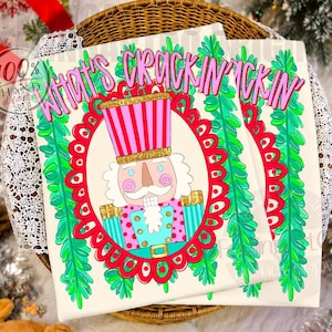 May include: A rectangular sign featuring a nutcracker illustration centred within a red scalloped oval frame. The nutcracker wears a pink and white striped hat with gold glitter accents. The text "WHAT'S CRACKIN'" appears in pink above the frame.